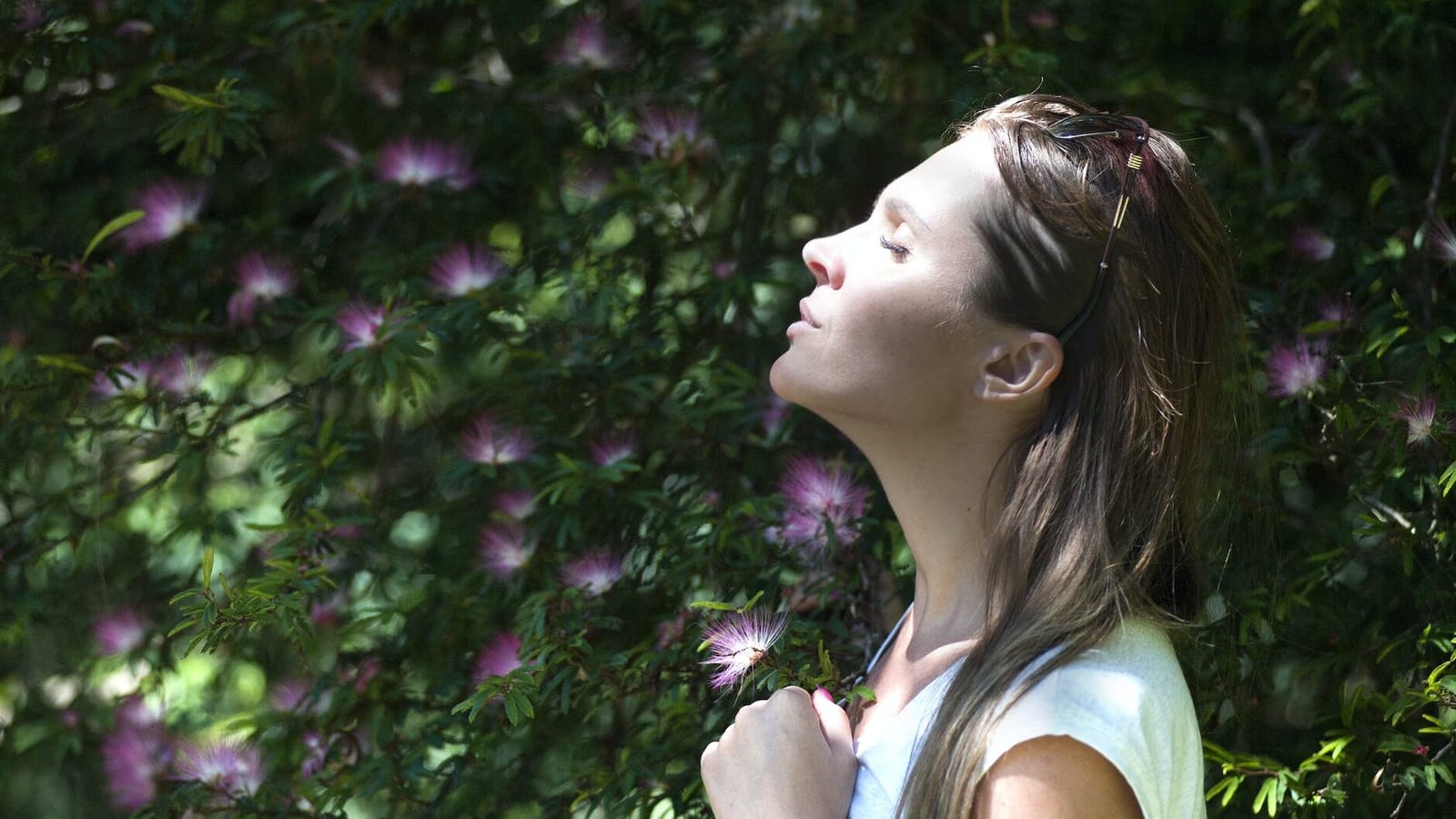 woman closing her eyes against sun light standing near purple petaled flower plant